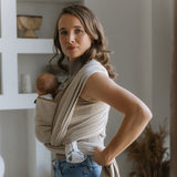 Baby wrapped in Tragetuch Leo Natur sling, soft diagonal weave, beige color, on a woman indoors.