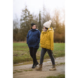 Two women outdoors wearing Mamalila Allwetter Tragejacke cosy allrounder jackets in blue and yellow with winter hats walking on a path in a natural setting