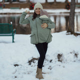 woman wearing green Mamalila Allwetter Tragejacke carrying baby in snowy outdoor park