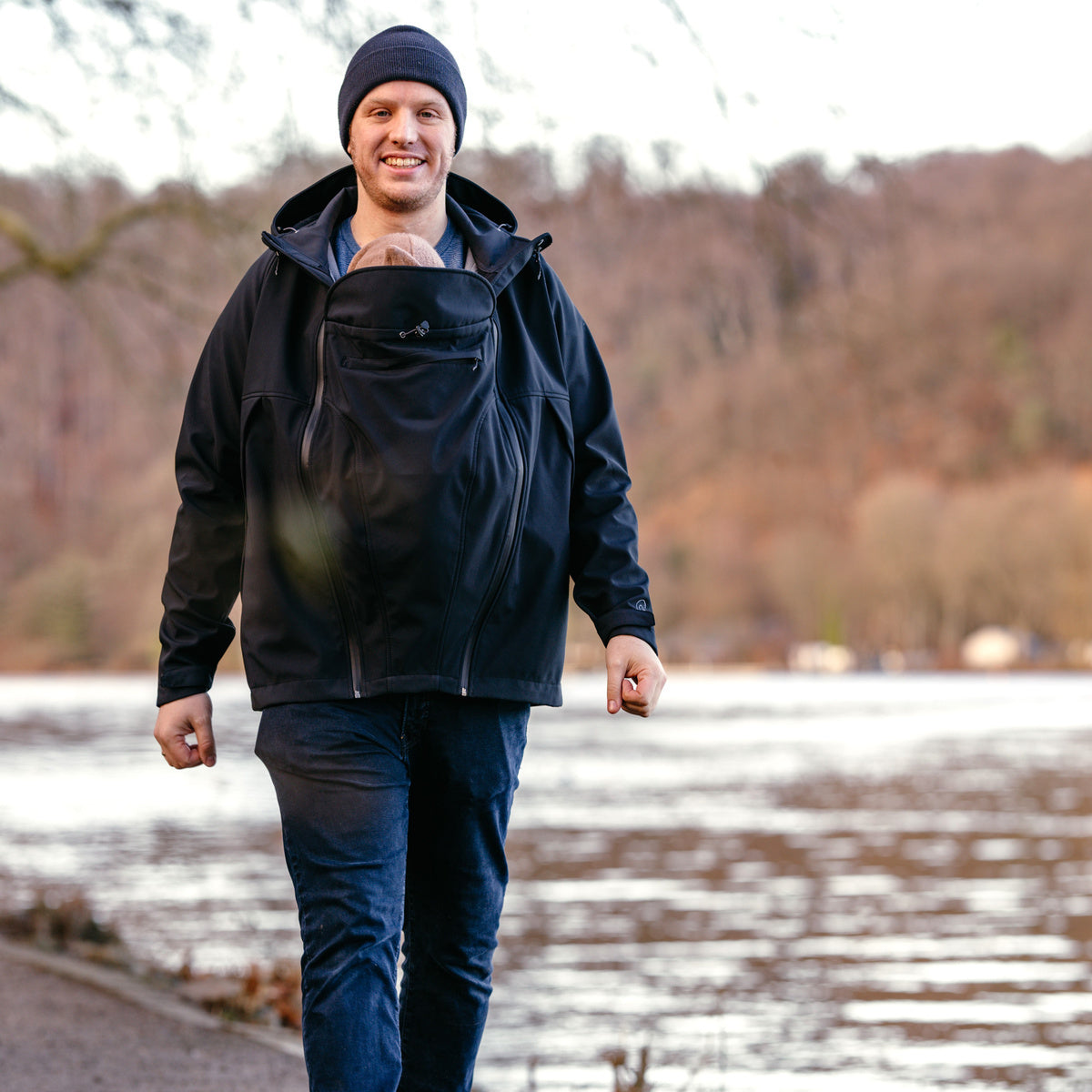 Man wearing Mamalila Softshell Tragejacke carrying baby in front pocket by lake outdoors